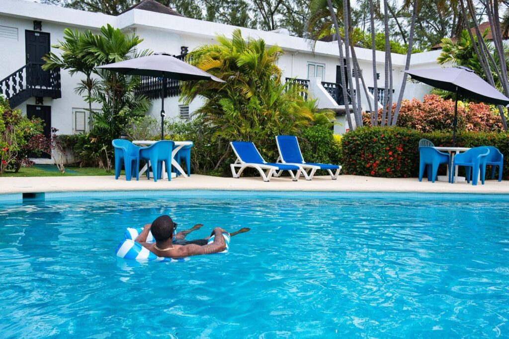 Guest floating in the swimming pool at Plum Tree Club Barbados, with apartment blocks and sun loungers behind